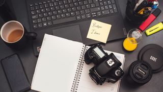 A workspace featuring a laptop, coffee cup, camera, notepad with "Budget Ideas," and colorful stationery on a dark surface
